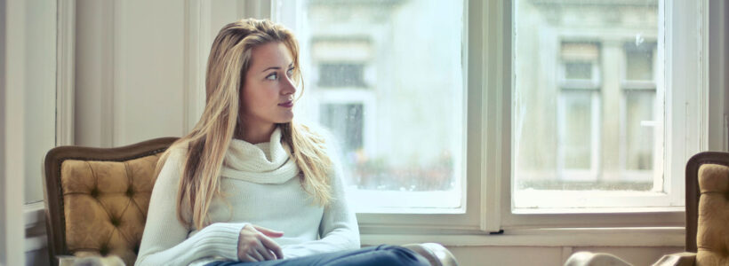 A woman is sitting on her couch in her apartment with a pensive expression on her face.