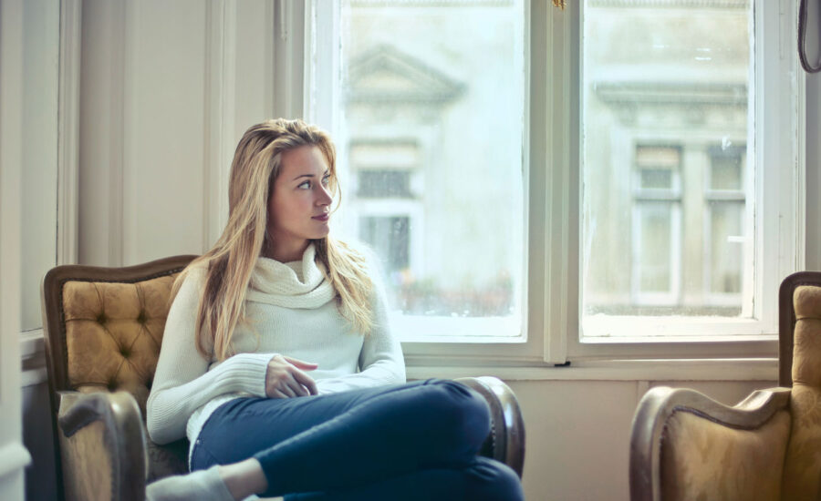 A woman is sitting on her couch in her apartment with a pensive expression on her face.