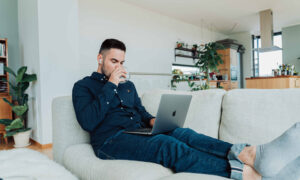 A man sits on his couch, feet up, sipping coffee, as he looks at his investments on his laptop.