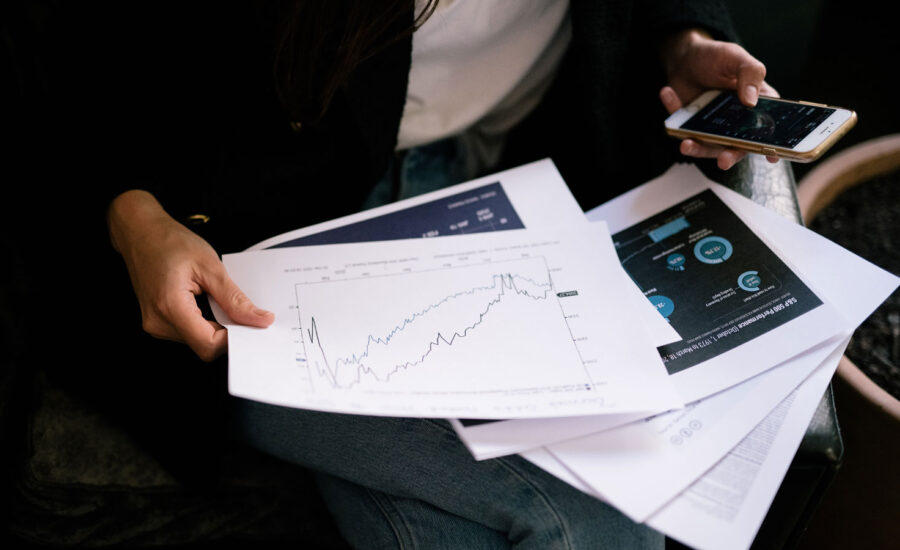 A woman is holding papers with graphs from the stock markets over the years, showing the dips. She also holds a cellphone in the other hand, implying that she may buy during inflation, bond and interest rates rising