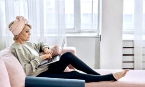 An older woman in yoga clothes sits on a sofa looking at a laptop.