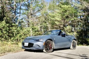 A grey Mazda MX-5 drives on a forest road