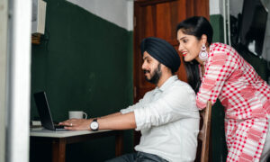 a man and a woman are sitting at a desk, smiling at what they're seeing on a laptop–as they research the value of certain stocks