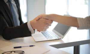 Two people shake hands across a desk that has a laptop on it