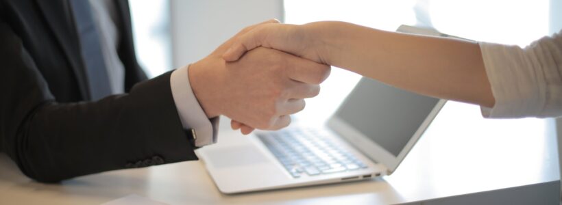 Two people shake hands across a desk that has a laptop on it