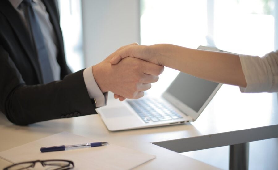 Two people shake hands across a desk that has a laptop on it