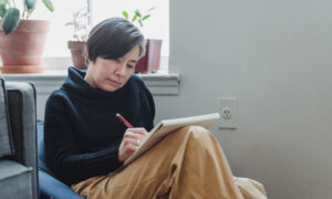 An older woman sits in a chair by the window, sketching with a pencil and paper.