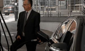 A man dressed for work is at the pump filling his car up with gas, thinking about inflation and the cost of gas as he returns to work.