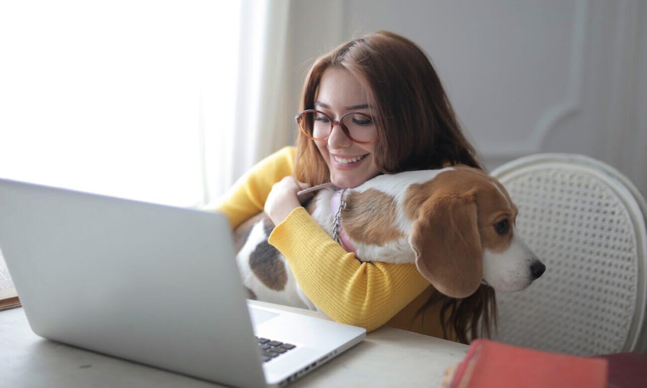 A young woman smiles at her laptop while hugging her dog