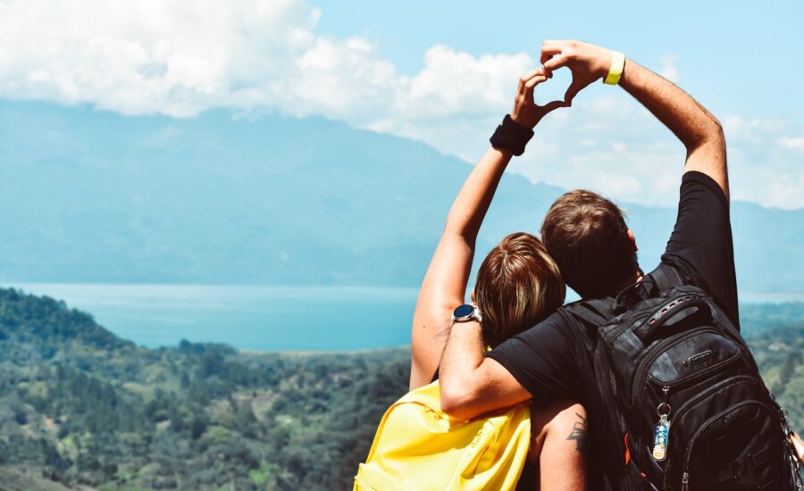 Two people with backpacks stand facing a mountain and lake, making a heart with their hands