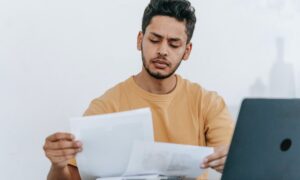 A man stairs intently at his auto insurance claim he received in the mail while his laptop remains open on a desk