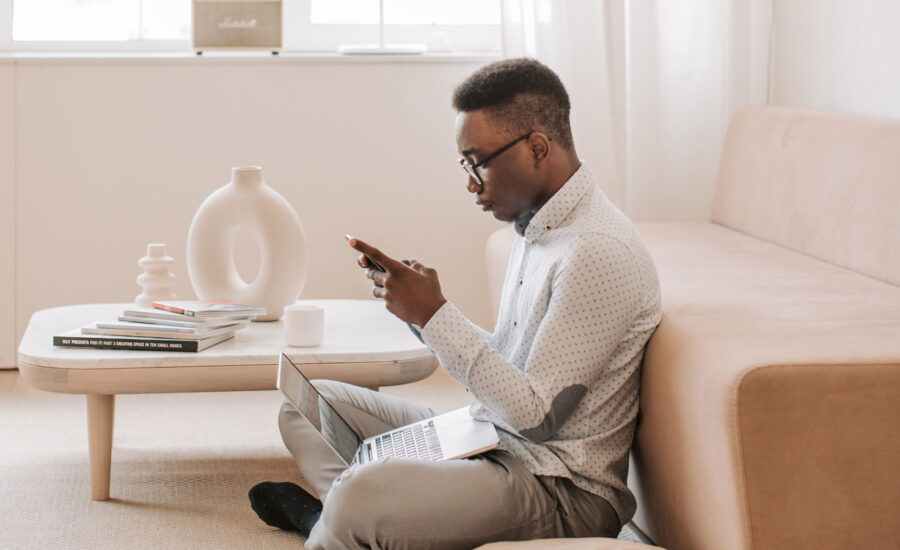 A Black man sits on his living room floor, checking both his laptop and phone app for cheap stock prices as he buys investment assets.