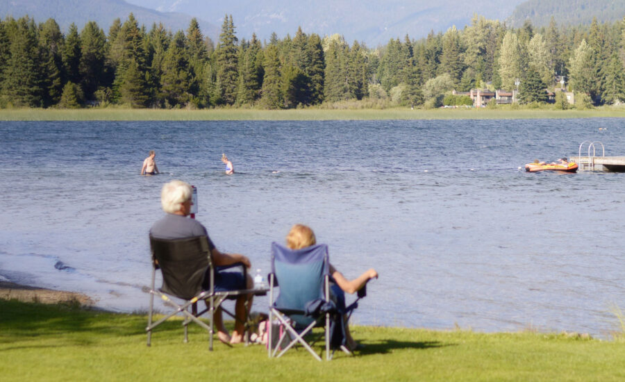 A couple is enjoying early retirement due to COVID, as they sit in the warmth of the sun at their cottage. They are cozy in camping chairs, facing a calm pond.