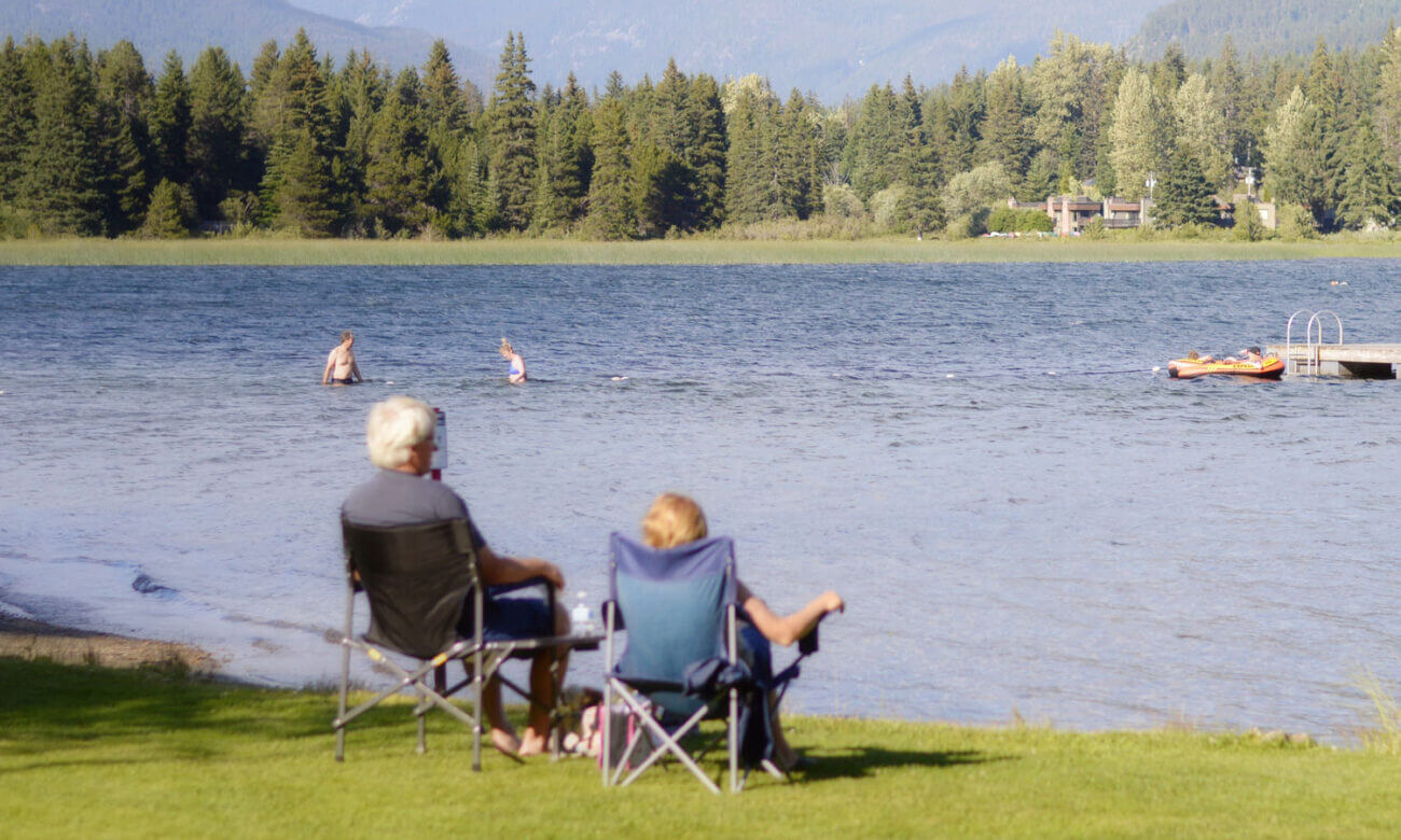 A couple is enjoying early retirement due to COVID, as they sit in the warmth of the sun at their cottage. They are cozy in camping chairs, facing a calm pond.