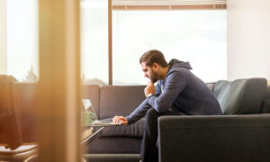 A young man wearing a hoodie, sitting on his couch is looking inquisitively at his laptop, wondering how he should remain a long-term investor within a bear market