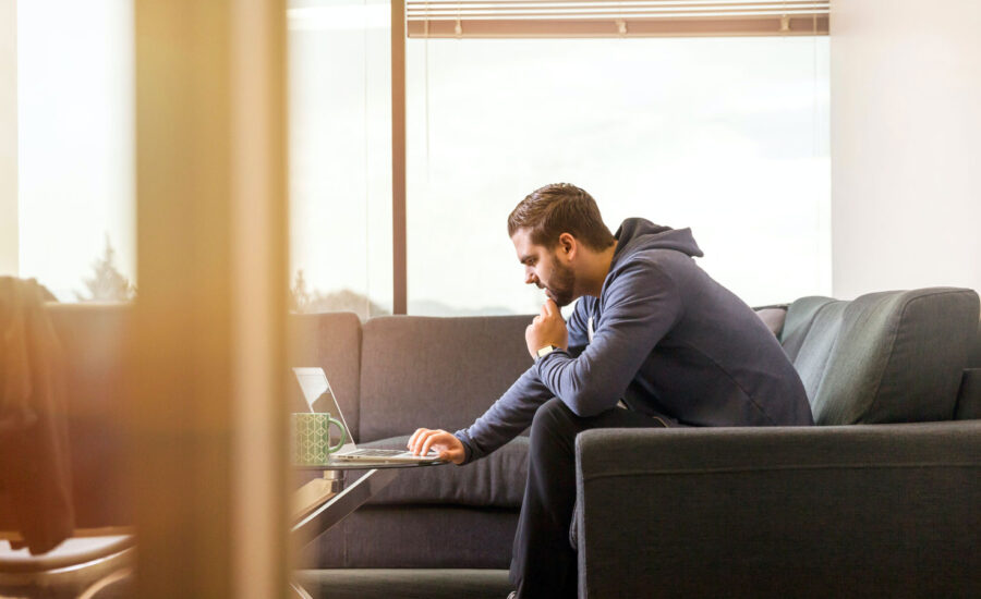 A young man wearing a hoodie, sitting on his couch is looking inquisitively at his laptop, wondering how he should remain a long-term investor within a bear market