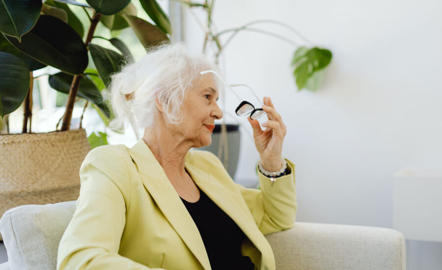 A woman in her 80s is sitting on her couch at home, removing her glasses, as she is thinking about selling her home or get a reverse mortgage.