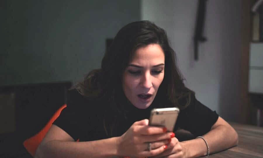 A woman stares at her phone in shock, realizing she's been scammed.