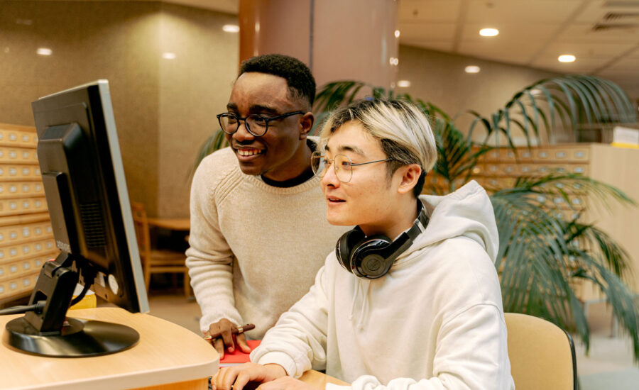 Two young men are in the library, using the computers as they're smiling. Why? Because they're not stressed about money and they've budgeted well.