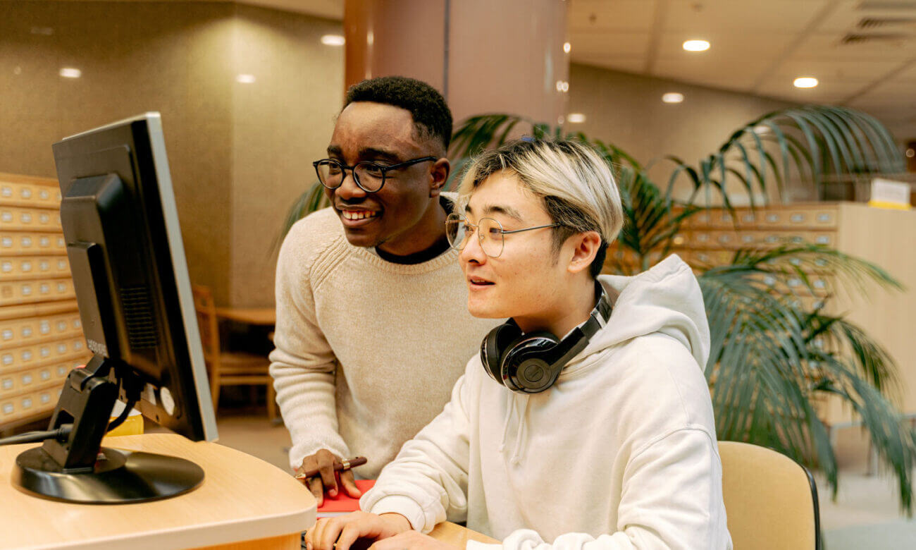 Two young men are in the library, using the computers as they're smiling. Why? Because they're not stressed about money and they've budgeted well.