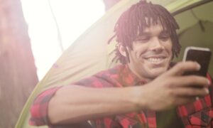 A man sits in front of a tent in the woods, smiling at his phone.