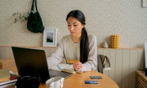 A woman sits at her computer, holding a credit card, contemplating using a line of credit to pay off her credit card debt