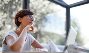 A woman sitting at her laptop gazes upward, deep in thought.