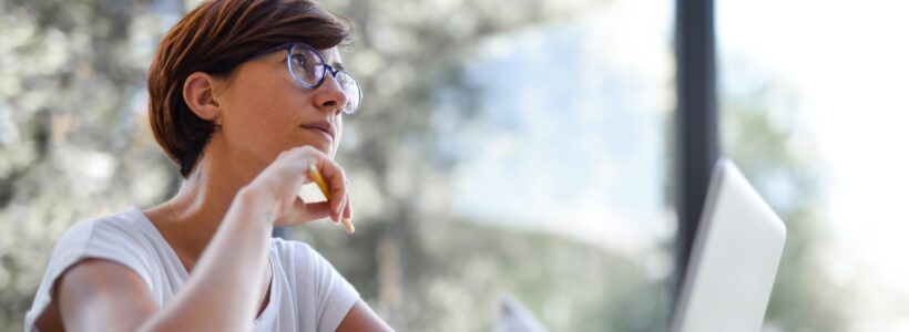 A woman sitting at her laptop gazes upward, deep in thought.