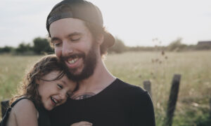 A man standing in a field considers whether he needs life insurance, while holding his young daughter in his arms