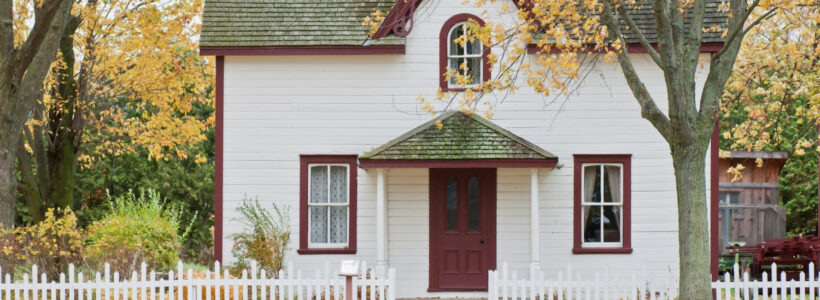 The streetview of a white house with red trim and a white picket fence in the fall