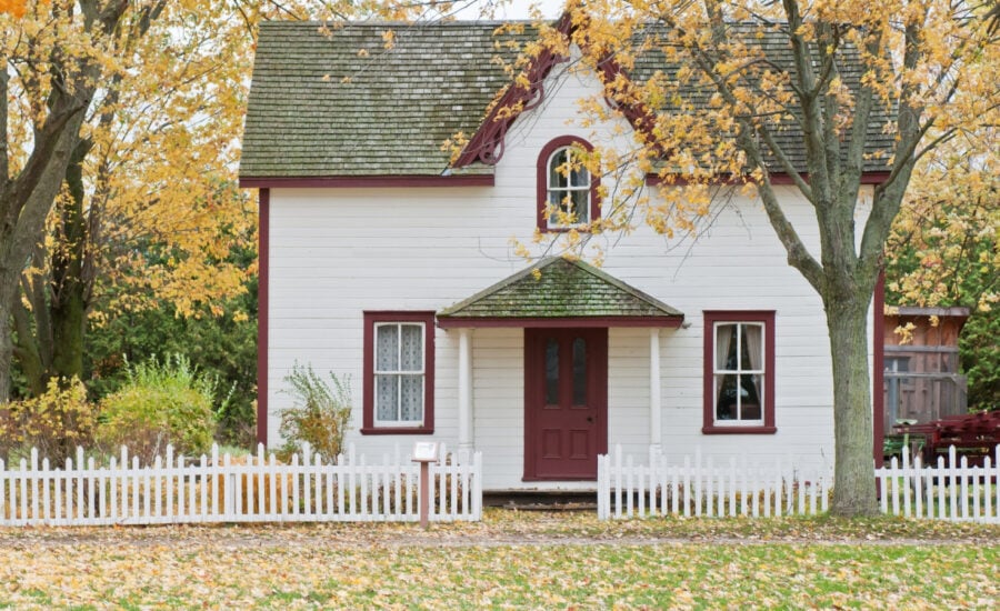 The streetview of a white house with red trim and a white picket fence in the fall