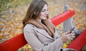 Young woman sits on a park bench smiling at her phone