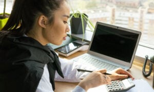 A woman is sitting at her desk, using her calculator to figure out her debt-to-income ratio.