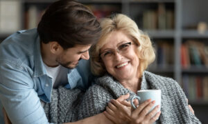 An adult man, acting as power of attorney, hugs his senior mom. He's wondering about gifting her assets while she's in a home.