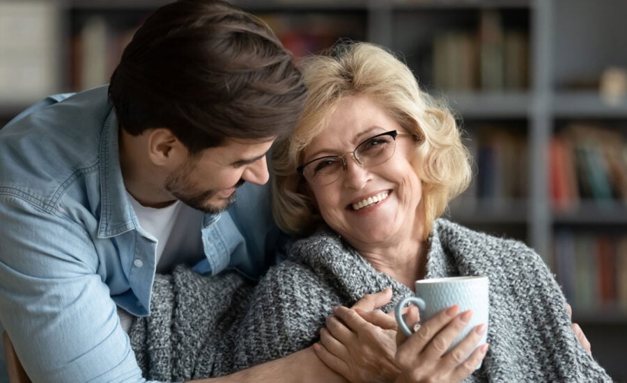 An adult man, acting as power of attorney, hugs his senior mom. He's wondering about gifting her assets while she's in a home.