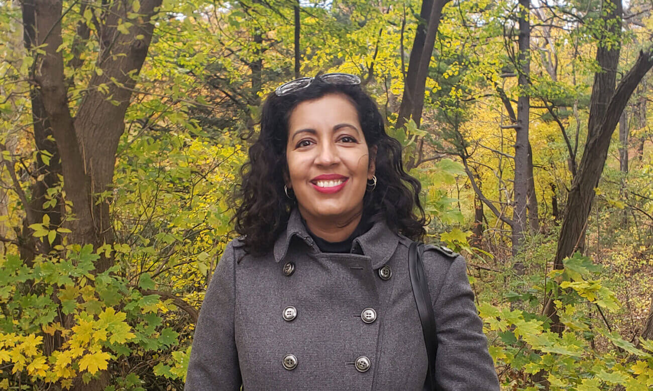 Renée Sylvestre-Williams smiles in a headshot of her taken on a beautiful fall day near some trees.
