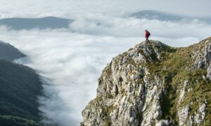 A person stands at the summit of a mountain, looking down at the clouds