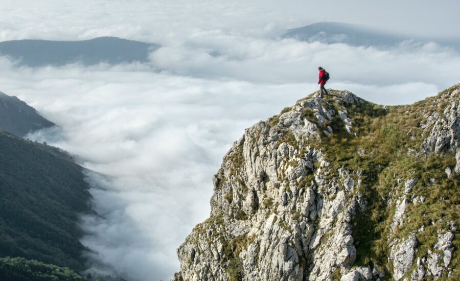 A person stands at the summit of a mountain, looking down at the clouds