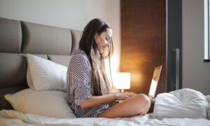 A smiling young woman sits on a hotel bed, looking at her laptop