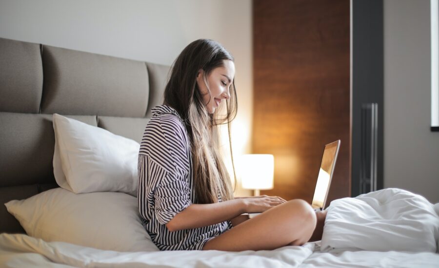 A smiling young woman sits on a hotel bed, looking at her laptop