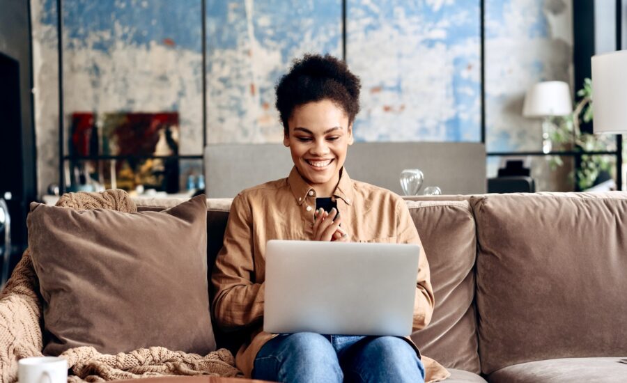A woman sits on her sofa and grins at her laptop.