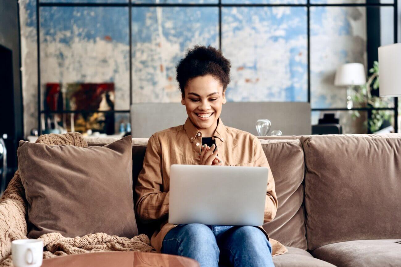 A woman sits on her sofa and grins at her laptop.