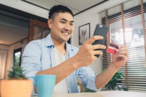 A smiling man looks at his phone while holding a credit card