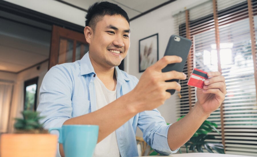 A smiling man looks at his phone while holding a credit card