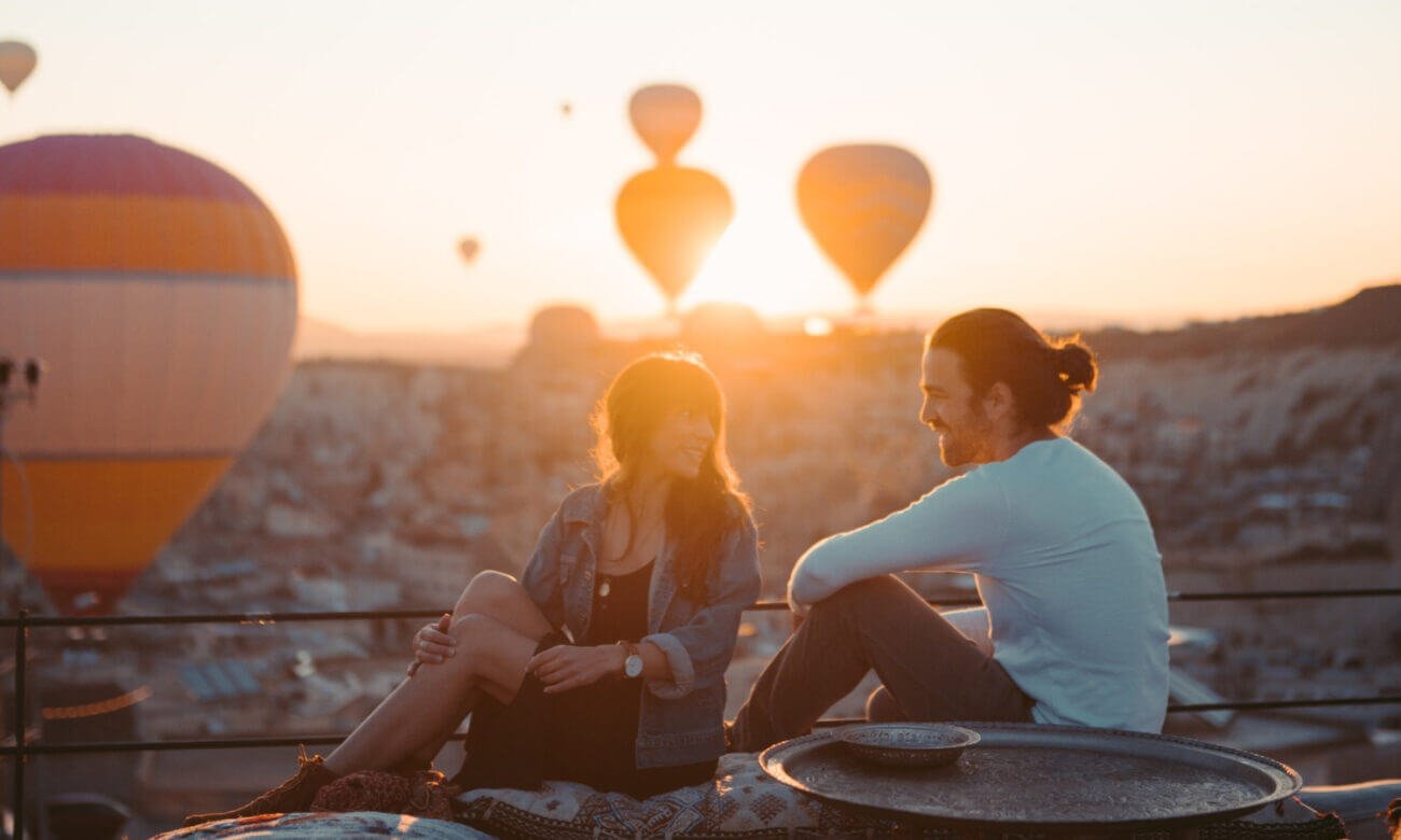 Friends sit together on a rooftop overlooking a sky filled with hot air balloons