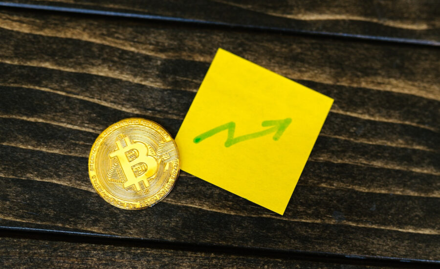 A gold coin with a Bitcoin logo sits beside a sticky note with an upward arrow