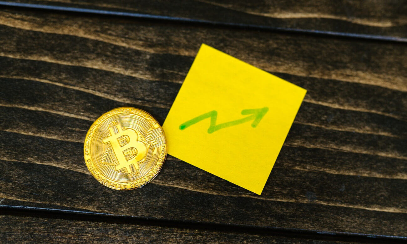 A gold coin with a Bitcoin logo sits beside a sticky note with an upward arrow