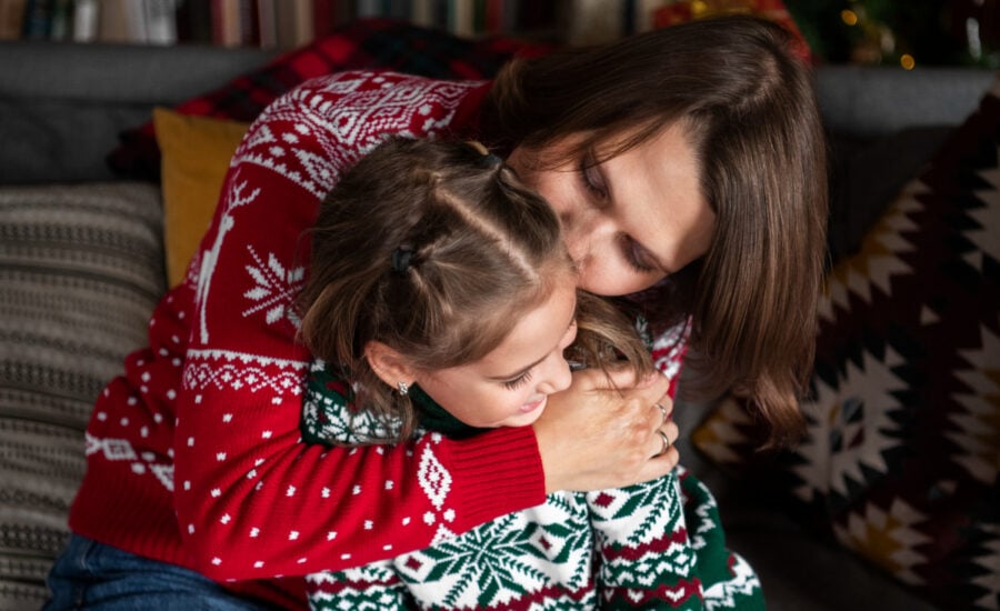 A woman hugs and kisses her young daughter, both wearing Christmas sweaters