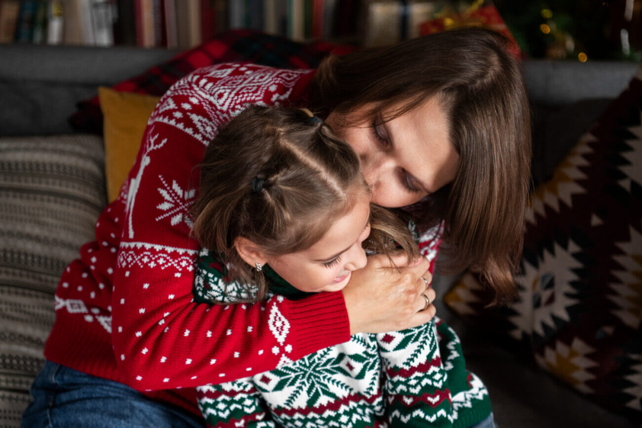 A woman hugs and kisses her young daughter, both wearing Christmas sweaters