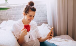 A woman drinks tea on a sofa, smiling at her phone